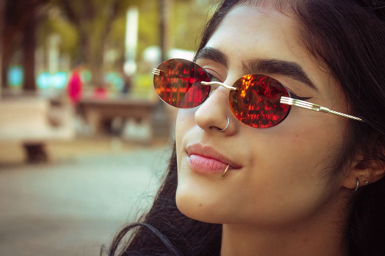 Close-up of a young woman with piercings wearing sunglasses in a stylish outdoor setting.