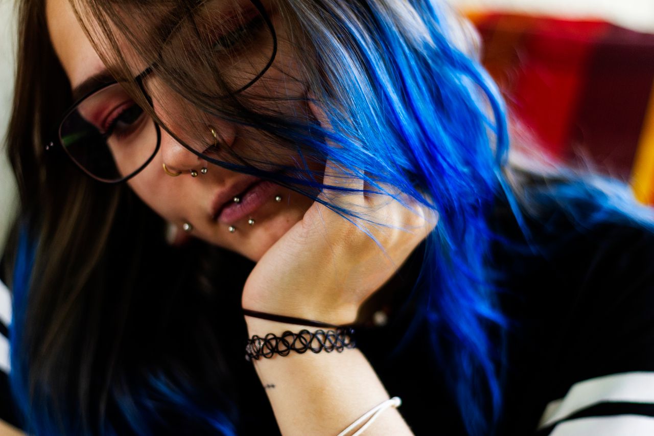 A close-up portrait of a young woman with vibrant blue hair, glasses, and multiple piercings.